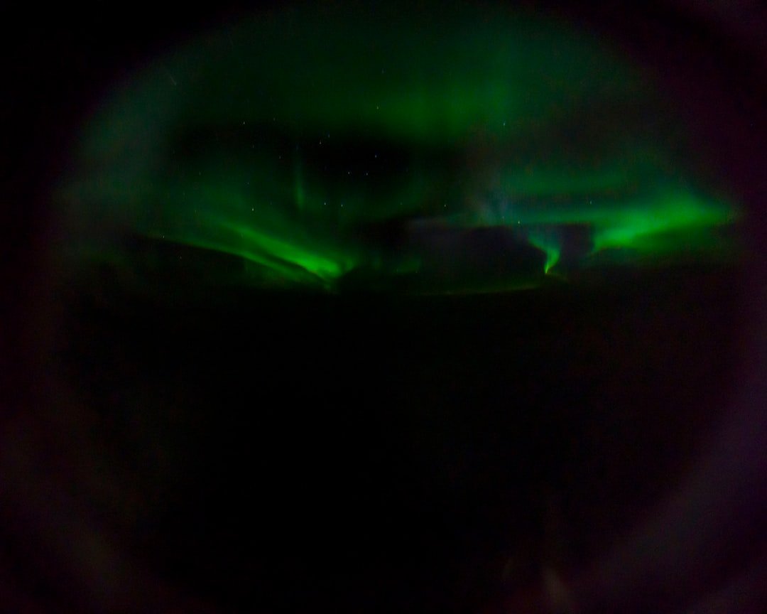 Photographer checking camera settings on tripod at night with faint green aurora visible on the horizon